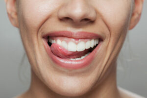 Close-up of a smiling woman with white teeth, licking her upper teeth with her tongue, showcasing healthy gums and a confident, clean smile.
